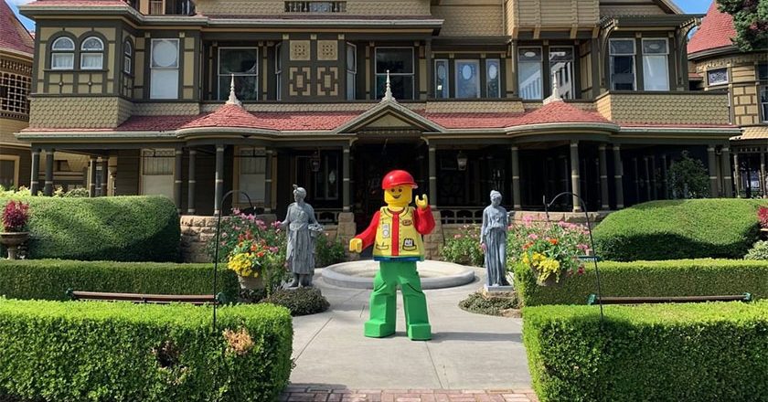 A costumed LEGO minifigure character standing in front of the Winchester Mystery House