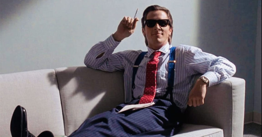 Christian Bale as Patrick Bateman sitting on a couch in sunglasses smoking a cigarette in "American Psycho."