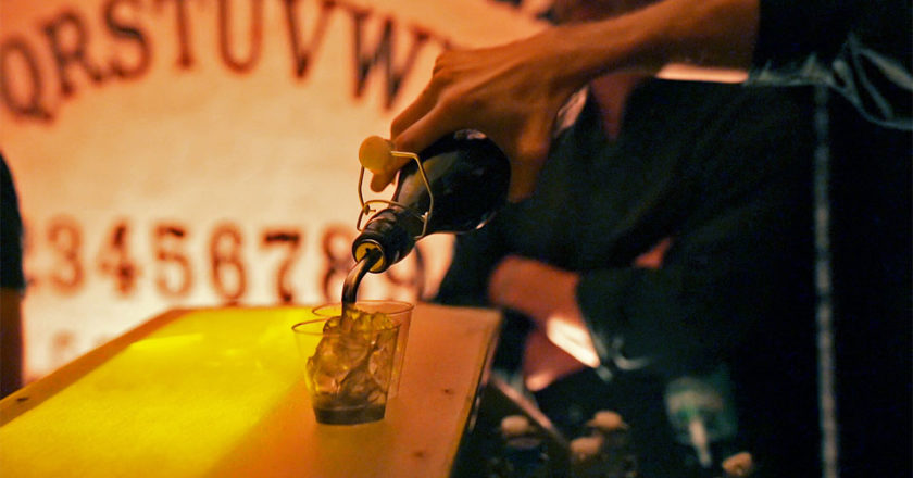 Bartender pouring a drink with a biant Ouija Board in the background.