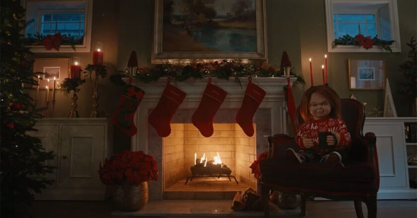 Chucky sits in a chair in front of a fireplace decorated for Christmas