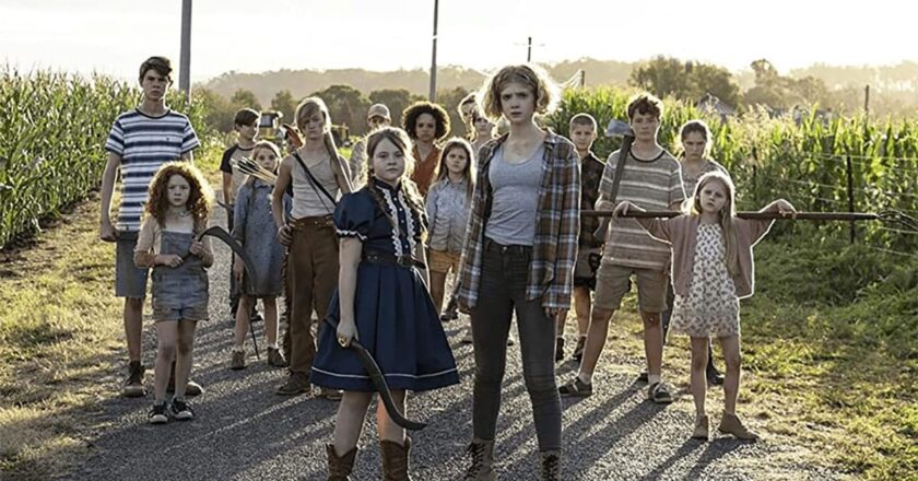 A group of children stand with farm tools on a dirt road between two corn fields