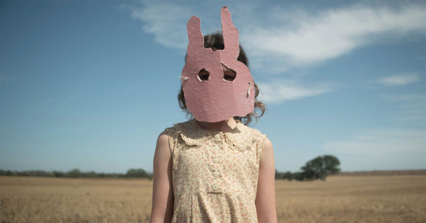 Still from "Run Rabbit Run" featuring a girl standing in a field wearing a pink rabbit mask made of cardboard.
