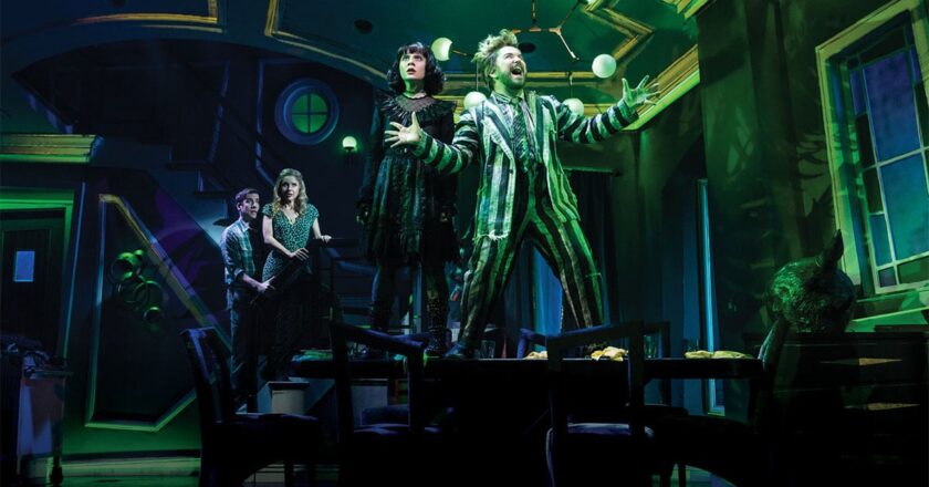 Lydia and Beetlejuice stand atop a table as Adam and Barbara look on from the stairs in Beetlejuice The Musical